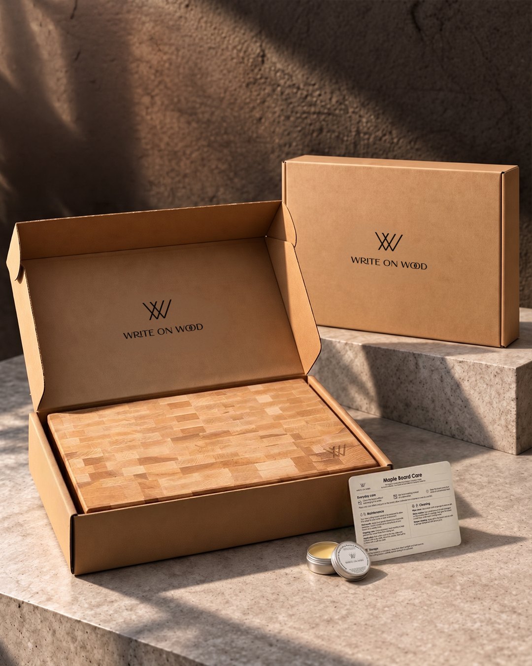 A hardwood board in an open Write On Wood gift box on stone, with a separate gift box, care card and beeswax tin